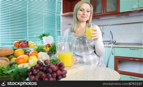 Pregnant woman drinking orange juice in the kitchen near a lot of vegetables and fruits