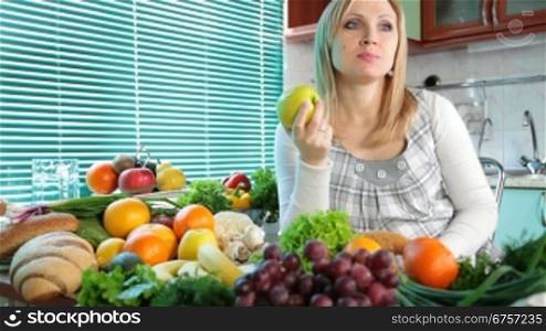 Pregnant woman eating apple near a lot of vegetables and fruits in the kitchen