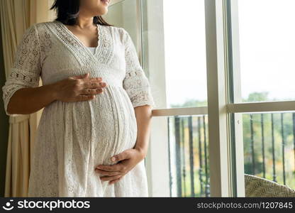 Pregnant woman feeling happy at home while taking care of her child. The young expecting mother holding baby in pregnant belly. Maternity prenatal care and woman pregnancy concept.