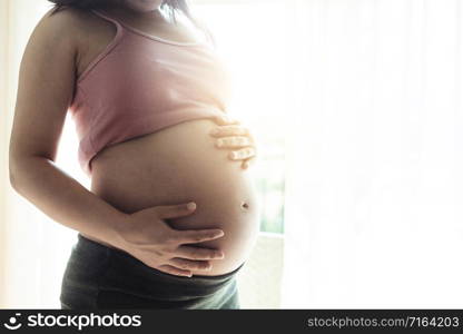 Pregnant woman feeling happy at home while taking care of her child. The young expecting mother holding baby in pregnant belly. Maternity prenatal care and woman pregnancy concept.