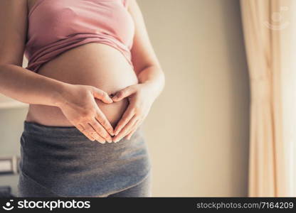 Pregnant woman feeling happy at home while taking care of her child. The young expecting mother holding baby in pregnant belly. Maternity prenatal care and woman pregnancy concept.