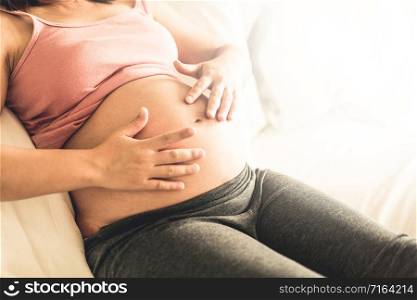 Pregnant woman feeling happy at home while taking care of her child. The young expecting mother holding baby in pregnant belly. Maternity prenatal care and woman pregnancy concept.