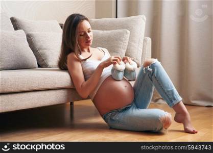 Pregnant woman holding small baby shoes relaxing at home by the couch
