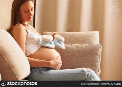 Pregnant woman holding small baby shoes relaxing at home on couch
