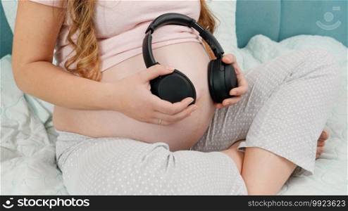 Pregnant woman in pajamas listening to music and holding headphones at her big belly. Unborn baby listening to classic music while being in mothers abdomen. Concept of early art and creativity development.. Pregnant woman in pajamas listening to music and holding headphones at her big belly. Unborn baby listening to classic music while being in mothers abdomen. Concept of early art and creativity development