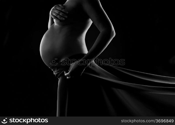 Pregnant woman in silk veil demonstrating her belly. Black and white studio shot on black background