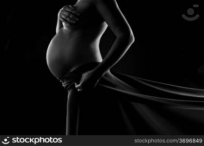 Pregnant woman in silk veil demonstrating her belly. Black and white studio shot on black background