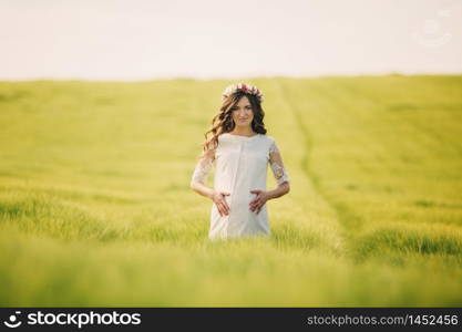 pregnant woman in white dress with wreath of flowers on head is keeping her hands on stomach and relaxing outdoors in grass field. motherhood concept, selective focus,. pregnant woman in white dress with wreath of flowers on head is keeping her hands on stomach and relaxing outdoors in grass field. motherhood concept, selective focus