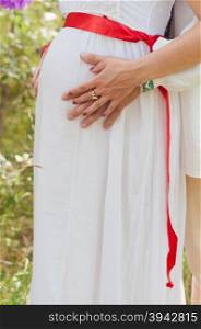 pregnant woman is touching her belly with hands. pregnant woman in white dress with red ribbon is touching her belly with hands