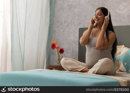 Pregnant woman listening to music with headphone while sitting on bed at home