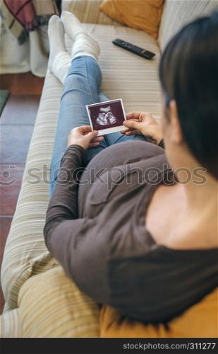Pregnant woman looking at the ultrasound of her baby in the living room. Pregnant looking ultrasound of her baby