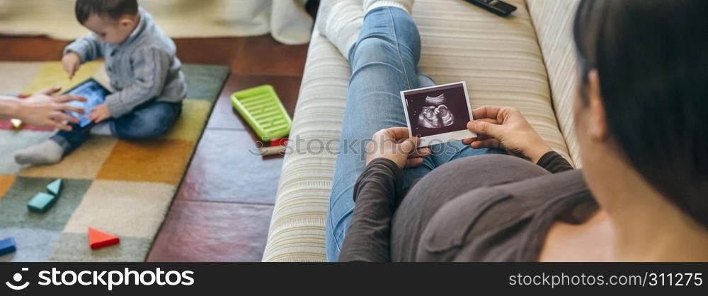 Pregnant woman looking at the ultrasound of her new baby while the older brother plays. Pregnant looking ultrasound of their new baby