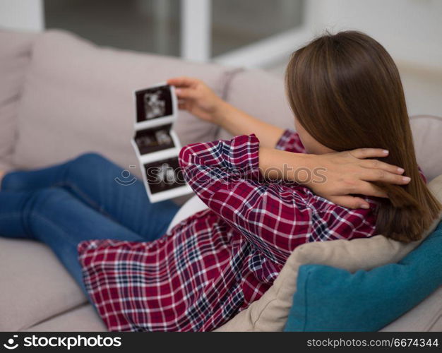 pregnant woman looking baby&rsquo;s ultrasound. Young pregnant woman looking baby&rsquo;s ultrasound photo while relaxing on sofa at home