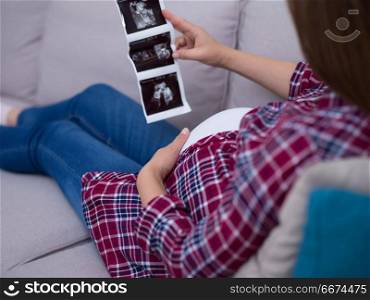 pregnant woman looking baby&rsquo;s ultrasound. Young pregnant woman looking baby&rsquo;s ultrasound photo while relaxing on sofa at home