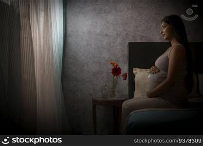 Pregnant woman looking out of window while sitting on bed at home