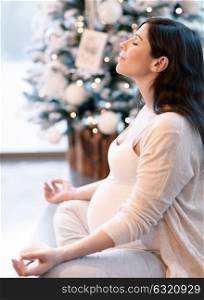 Pregnant woman meditating at home near beautiful decorated Christmas tree, sitting in lotus pose with closed eyes, body care and inner peace