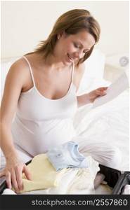 Pregnant woman packing baby clothing in suitcase holding list smiling