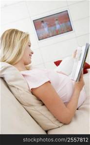 Pregnant woman reading magazine with television in background smiling