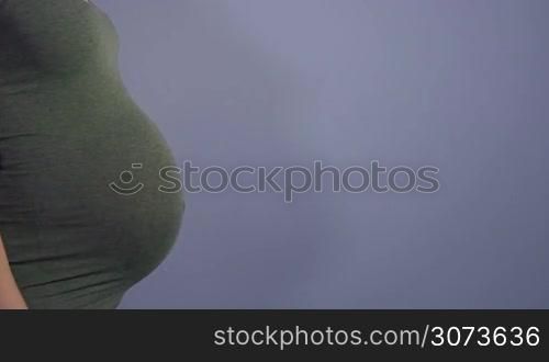 Pregnant woman showing belly and female hands touching it, mom with expectant tummy on blue background, mother during pregnancy. Maternity, motherhood, new life and fertility. Copy space, slow motion