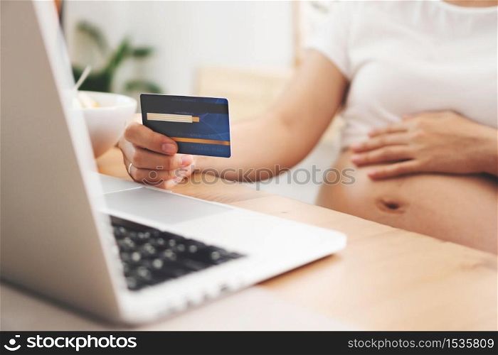 Pregnant Woman sitting holding blue credit card and making online payment shopping on laptop computer at home. Female making purchases in the Internet banking. Spending money and lifestyle.