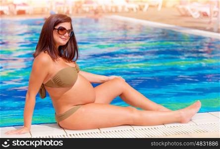 Pregnant woman sitting near the pool, expectant female relaxing at luxury summer resort, healthy lifestyle, happy pregnancy concept