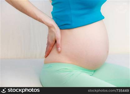 Pregnant woman sitting on sofa at home with hands on her back, making massage, suffering from pain. Pregnancy and health concept. Pregnant woman sitting on couch holds hand on her back