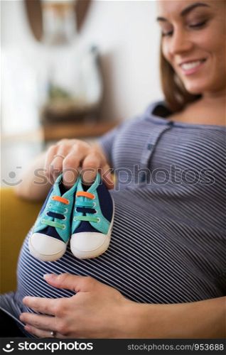 Pregnant Woman Sittingh On Sofa At Home Holding Baby Shoes