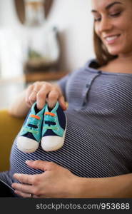 Pregnant Woman Sittingh On Sofa At Home Holding Baby Shoes