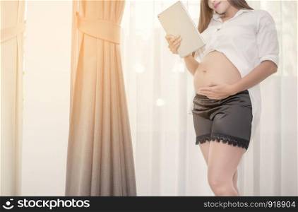 Pregnant woman standing and using the tablet with touch her bellow in her house, Family and Health care concept