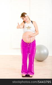 Pregnant woman standing on weight scale with measure tape and showing thumbs up &#xA;