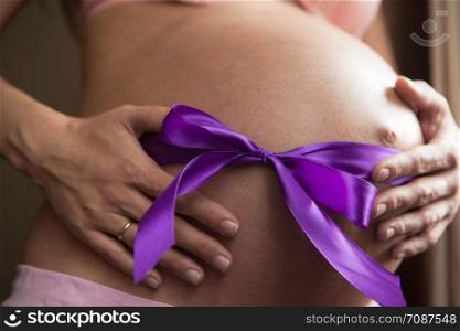 Pregnant woman supports her belly. A purple ribbon is tied to the bow on the belly. Concept of pregnancy as a gift, happiness of parenthood. Lifestyle photo. Horizontal orientation of the frame.