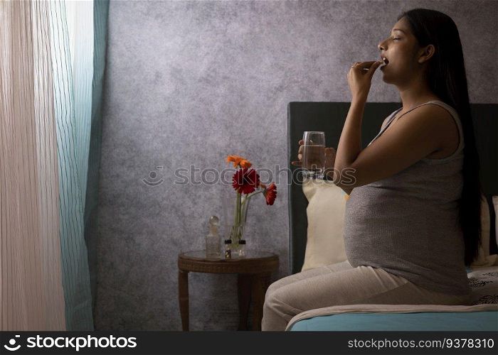 Pregnant woman talking medicine while sitting on bed at home