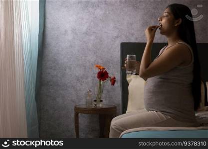 Pregnant woman talking medicine while sitting on bed at home