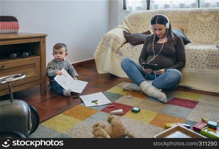 Pregnant woman using the tablet sitting on the carpet in the living room while her son is playing. Pregnant using the tablet while her son is playing