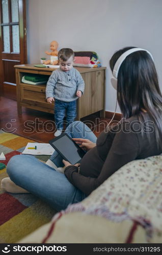 Pregnant woman using the tablet sitting on the carpet in the living room while her son is playing. Pregnant using the tablet while her son is playing