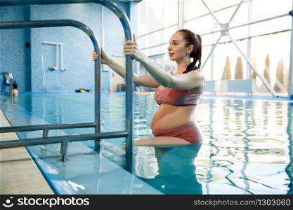 Pregnant woman with big belly climbs a ladder out of the water in the pool indoors. Pregnancy being, relaxation in the water, recreational exercise for health care. Pregnant woman climbs a ladder out of the water
