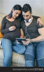 Pregnant woman with her partner looking at the tablet and eating chocolate. Pregnant with her partner looking at the tablet