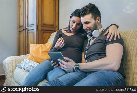 Pregnant woman with her partner looking at the tablet and eating chocolate. Pregnant with her partner looking at the tablet