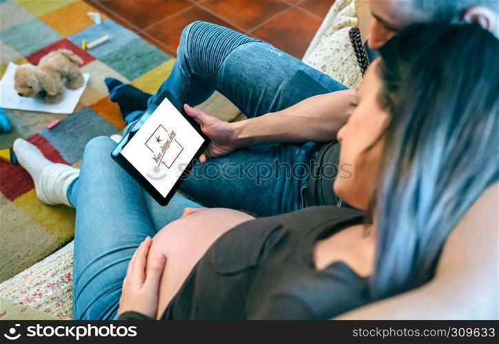 Pregnant woman with her partner looking in the tablet a ultrasound of their baby in the living room. Customizable tablet screen. Pregnant with her partner looking ultrasound of their baby