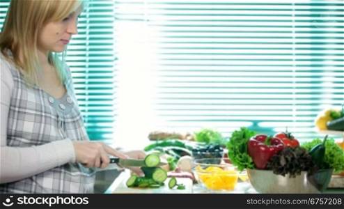 Pregnant woman with knife cutting cucumber on board in kitchen
