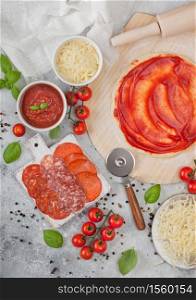 Preparation for baking of pepperoni pizza with raw dough, salami spicy chorizo with wheel cutter and fresh tomatoes and basil on light table with bowl plates with cheese and tomato paste.