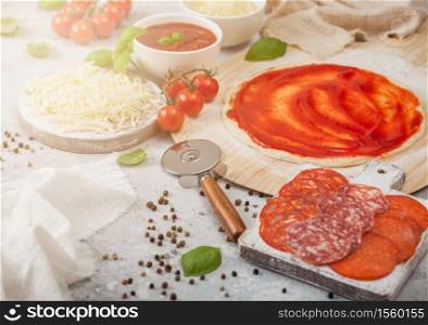 Preparation for baking of pepperoni pizza with raw dough, salami spicy chorizo with wheel cutter and fresh tomatoes and basil on light table with bowl plates with cheese and tomato paste.