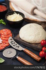 Preparation for baking of pepperoni pizza with raw dough, salami spicy chorizo with wheel cutter and fresh tomatoes and basil on dark table with bowl plates with cheese and tomato paste.