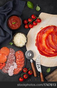 Preparation for baking of pepperoni pizza with raw dough, salami spicy chorizo with wheel cutter and fresh tomatoes and basil on dark table with bowl plates with cheese and tomato paste.