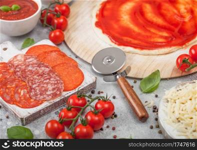 Preparation for baking of pepperoni pizza with raw dough, salami spicy chorizo with wheel cutter and fresh tomatoes and basil on light yable with bowl plates with cheese and tomato paste.