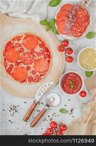 Preparation for baking of pepperoni pizza with salami spicy chorizo with wheel cutter and fresh tomatoes and basil on light table with bowl plates with cheese and tomato paste.