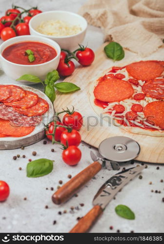 Preparation for baking of pepperoni pizza with salami spicy chorizo with wheel cutter and fresh tomatoes and basil on light table with bowl plates with cheese and tomato paste.