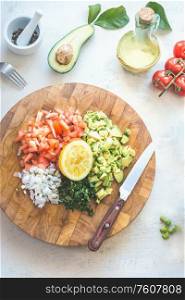 Preparation of tasty fresh avocado tomato salsa salad on white kitchen table background, top view. Chopped vegetables on wooden cutting board. Healthy food. Vegetarian lunch or snack