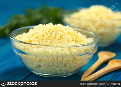 Prepared couscous in glass bowl on blue wooden surface with parsley in the back (Selective Focus, Focus one third into the couscous). Prepared Couscous in Glass Bowl