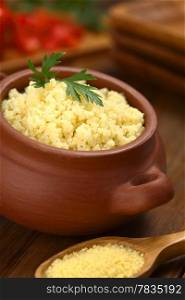 Prepared couscous in rustic bowl garnished with parsley, raw couscous on wooden spoon in front (Selective Focus, Focus on the front of the parsley leaf on the couscous). Prepared Couscous in Rustic Bowl